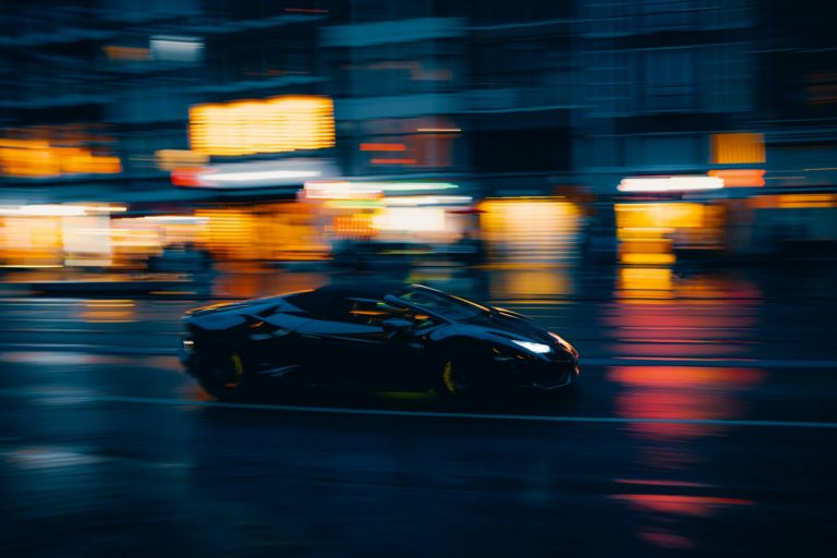A dark car speeds through a blurry city at night.